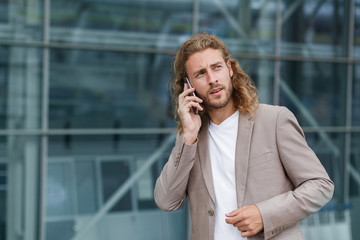 Young businessman talking on the phone, standing on city street near business center. Successful manager makes a call. Portrait of handsome curly smiling man in casual wear holding smartphone.