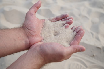 A man's hands with white sand. Sand falls through the gap between the palms, forming a funnel. There is also white sand in the background