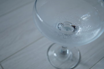 The cocktail glass stands on a light wooden plank surface. A glass in the right part of the frame, with sparkling water in it