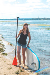Beautiful millennial woman with a SUP board and puddle on the river. Active rest concept. Standing on a coast
