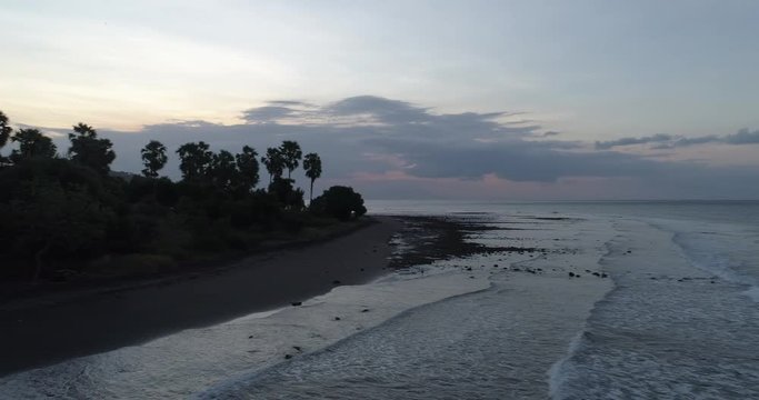 Drone shot at low altitude flying backwards over waves breaking on black sandy beach at dusk