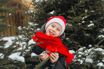 A teenage European boy in a red Santa Claus costume holds a Christmas gift in his hands . A child stands by a green fir tree. The boy is smiling and happy that it 's time for Christmas and New year.