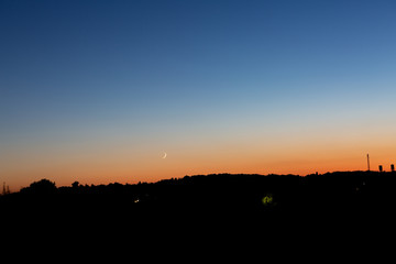 A young moon on an orange-blue sunset sky over a dark forest. Horizontal orientation. High quality photo