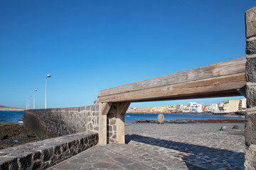 Fototapeta premium Views of El Medano town framed by a narrow wooden gangway and cobblestone pathways, towards the bay and residences behind, hotels and restaurants, beaches and coast, in Tenerife, Canary Islands, Spain