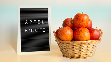 Ripe red and orange apples in wicker basket with handles on right and white word in german meaning Apples discounts on letter board on gradient yellow-blue backdrop,natural light and shadow.Copy space