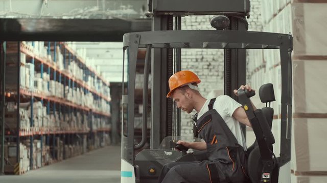 A seriou worker is sitting in a forklift car. He i settng the machine to work. The man is wearing a helmet and uniform.