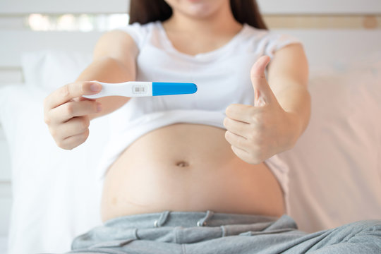 Closeup front view. Beautiful pregnant woman sitting on bed holding pregnancy test with two stripes confirm that female is pregnant. Pregnancy, motherhood, love and happy concept.