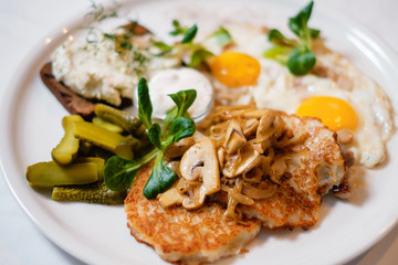 Close-up potato pancakes with mushrooms and fried eggs, served with bread and pickled cucumbers. The concept of a delicious hearty homemade breakfast served in a restaurant.
