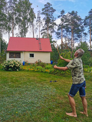 A tanned adult man plays badminton barefoot on the grass in the afternoon against the backdrop of a country house, tall pine trees and a blue sky with clouds.