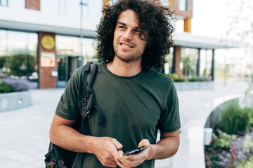 Confident man with curly hair wearing green t-shirt walking in the city street. Happy student male has joyful expression while resting outside.