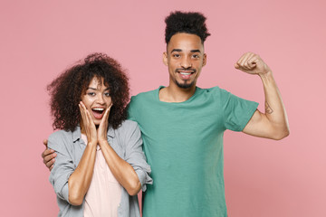 Amazed young african american couple friends guy girl in gray green casual clothes posing hugging showing biceps muscles put hands on cheeks isolated on pastel pink color background studio portrait.