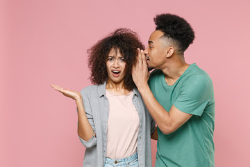 Shocked perplexed young african american couple friends guy girl in gray green casual clothes whispering secret behind her hand, sharing news isolated on pastel pink color background studio portrait.