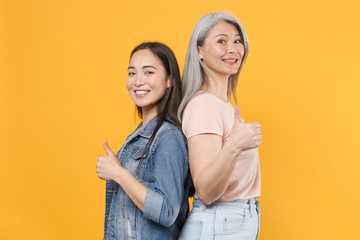 Smiling family asian female women girls gray-haired mother and brunette daughter in casual clothes posing standing back to back showing thumbs up isolated on yellow color background studio portrait.