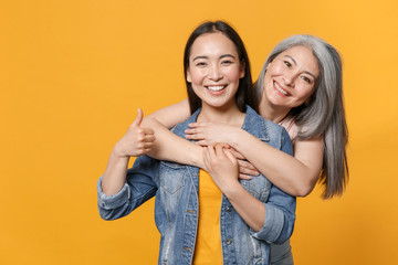 Smiling cheerful funny family asian female women girls gray-haired mother and brunette daughter in casual clothes posing hugging showing thumb up isolated on yellow color background studio portrait.