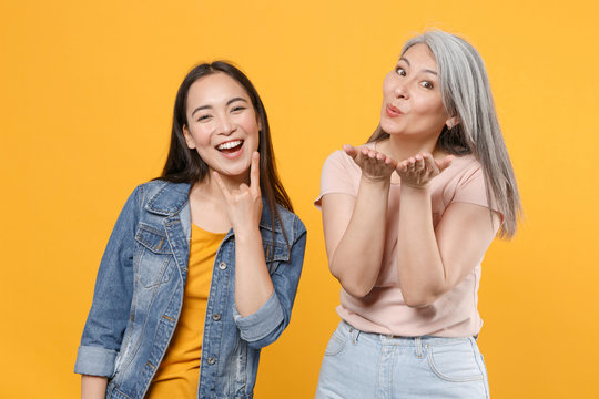 Funny family asian women girls gray-haired mother brunette daughter in casual clothes depicting heavy metal rock sign blowing sending air kiss isolated on yellow color wall background studio portrait.