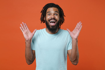 Surprised young african american man guy wearing blue casual t-shirt posing isolated on orange wall background studio portrait. People emotions lifestyle concept. Keeping mouth open, spreading hands.