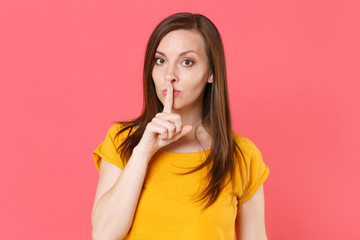 Secret young brunette woman 20s wearing yellow casual t-shirt posing standing saying hush be quiet with finger on lips shhh gesture looking camera isolated on pink color background studio portrait.