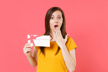 Shocked amazed young brunette woman 20s in yellow casual t-shirt posing holding in hand gift certificate covering mouth with hand looking camera isolated on pink color wall background studio portrait.