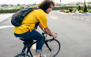 Outdoor shot of a young man in yellow shirt cycling on his bike with backapack in the city street. Male courier with curly hair delivers parcel cycling with a bicycle in the city.