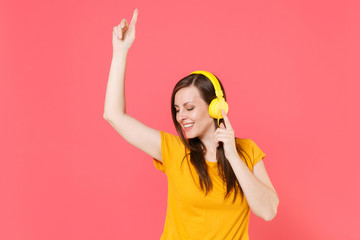Funny smiling young brunette woman 20s wearing yellow casual t-shirt posing listen music with headphones dancing rising hand pointing index finger up isolated on pink color background studio portrait.