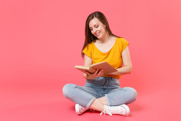 Full length portrait of smiling beautiful attractive young brunette woman 20s in yellow casual t-shirt posing sitting on floor and reading book isolated on pink color wall background studio.