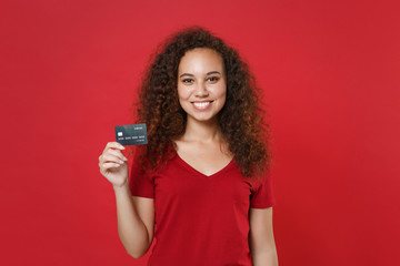 Smiling young african american woman girl in casual t-shirt posing isolated on red background studio portrait. People sincere emotions lifestyle concept. Mock up copy space. Hold credit bank card.