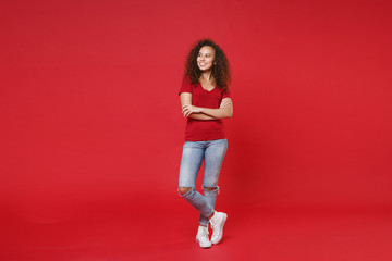 Full length portrait smiling young african american woman girl in casual t-shirt posing isolated on red background. People lifestyle concept. Mock up copy space. Holding hands crossed, looking aside.