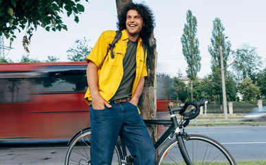 Young man resting after cycling on his bike in the city street. Male student with curly hair in yellow shirt with a bicycle waiting his friends outside.