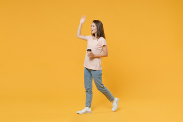 Full length side view portrait of smiling young woman in pink casual t-shirt hold paper cup of coffee or tea waving greeting with hand as notices someone isolated on yellow color background studio.