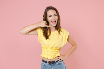 Blinking funny young brunette woman 20s wearing yellow casual t-shirt posing doing phone gesture like says call me back looking camera isolated on pastel pink color wall background studio portrait.