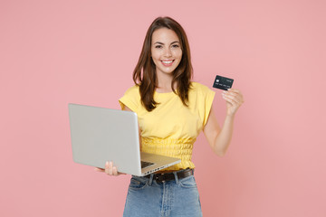Naklejka premium Smiling young brunette woman wearing yellow casual t-shirt posing standing working on laptop pc computer hold credit bank card looking camera isolated on pastel pink color background studio portrait.