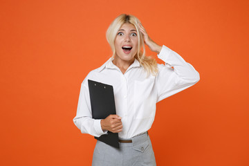 Shocked young blonde female business woman girl wearing white shirt posing hold clipboard with papers document put hand on head keeping mouth open isolated on orange color background studio portrait.