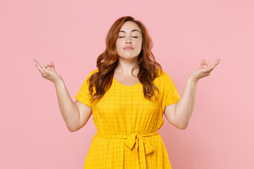 Young redhead plus size body positive female woman girl in yellow dress hold hands in yoga gesture, relaxing meditating, trying to calm down isolated on pastel pink color background studio portrait.