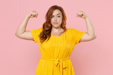 Obraz na płótnie Canvas Strong perplexed young redhead plus size body positive female woman girl 20s in yellow dress posing showing biceps, muscles looking camera isolated on pastel pink color background studio portrait.