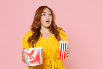 Shocked young redhead plus size body positive female woman girl in yellow dress watching movie film hold bucket of popcorn, cup of soda looking aside isolated on pink color background studio portrait.