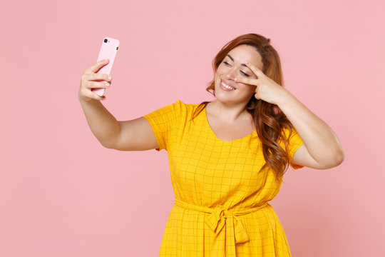 Funny young redhead plus size body positive female woman girl in yellow dress posing doing selfie shot on mobile phone showing victory sign isolated on pastel pink color background studio portrait.
