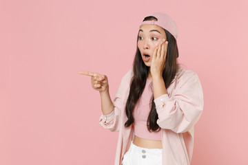 Shocked young asian woman girl in casual clothes cap posing isolated on pastel pink wall background studio. People lifestyle concept. Mock up copy space. Pointing index finger aside put hand on cheek.