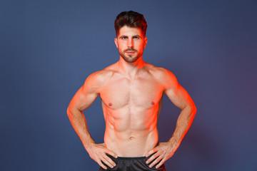 Fototapeta premium Handsome young bearded fitness sporty strong guy bare-chested muscular sportsman isolated on blue background studio. Workout sport motivation lifestyle concept. Standing with arms akimbo on waist.
