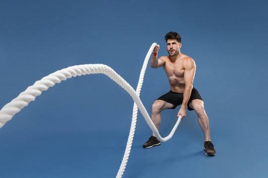 Full length portrait of young bearded fitness sporty strong guy bare-chested muscular sportsman isolated on blue background. Workout sport motivation lifestyle concept. Working out with battle ropes.