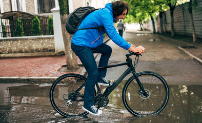 Side view image of young man cycling on his bike down the street next to the house. Male courier with curly hair in blue raincoat delivers parcel cycling with a bicycle in the city in a rainy day.