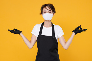 Young woman 20s barista bartender barman employee in apron sterile face mask to safe from coronavirus virus covid-19 2019-ncov during quarantine hold hands yoga gesture isolated on yellow background.