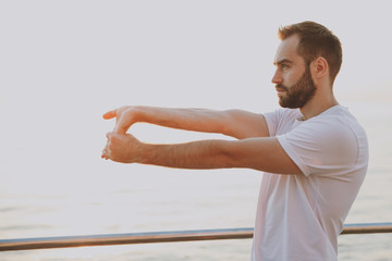 Side view portrait of young handsome bearded athletic man guy 20s in casual white t-shirt posing training doing stretching exercising warming up looking aside at sunrise over the sea outdoors.