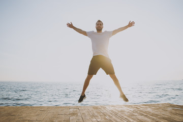 Fototapeta na wymiar Side view full length portrait of strong young bearded athletic man guy 20s in casual white t-shirt black shorts posing training jumping spreading hands and legs at sunrise over the sea outdoors.