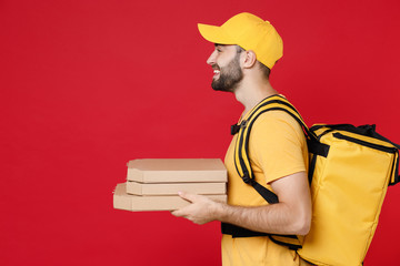 Side view delivery man in yellow cap t-shirt thermal bag backpack with food hold pizza in cardboard flatbox isolated on red background studio Employee work courier Service coronavirus covid-19 concept