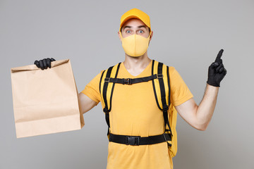 Delivery employee man in yellow cap face mask gloves tshirt thermal backpack hold craft paper takeaway food bag work courier service during quarantine covid-19 virus isolated on grey background studio