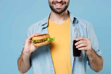 Cropped image of smiling young bearded man guy 20s wearing casual clothes posing holding american classic fast food burger bottle of cola isolated on pastel blue color wall background studio portrait.