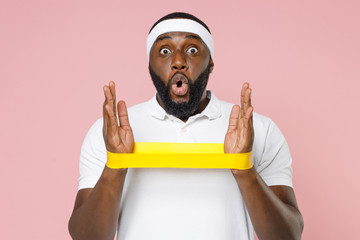 Shocked young bearded african american fitness sports man 20s in white headband t-shirt doing exercise with fitness gums spending time in gym isolated on pastel pink color background studio portrait.