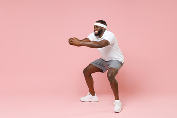 Full length portrait of smiling tired young bearded african american fitness sports man 20s in headband t-shirt doing exercise squatting spending time in gym isolated on pink color background studio.