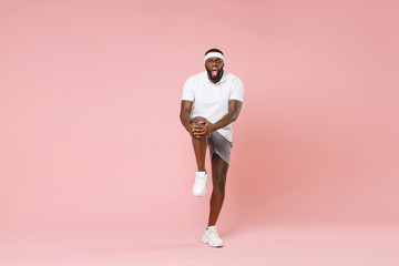Fototapeta na wymiar Full length portrait of displeased young african american fitness sports man 20s in headband t-shirt doing stretching exercising for legs spending time in gym isolated on pink color background studio.