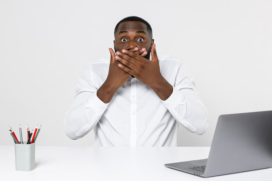 Shocked young african american male business man in white shirt posing working in office sitting at desk with laptop pc covering mouth with hands looking camera isolated on white color background.